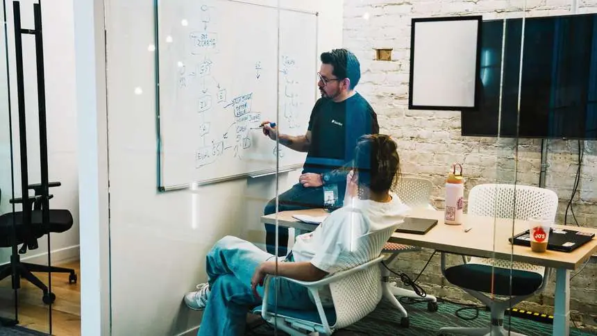 An instructor sketching an evaluation pipeline on a whiteboard with a Challenger in a glass meeting room at Gauntlet HQ